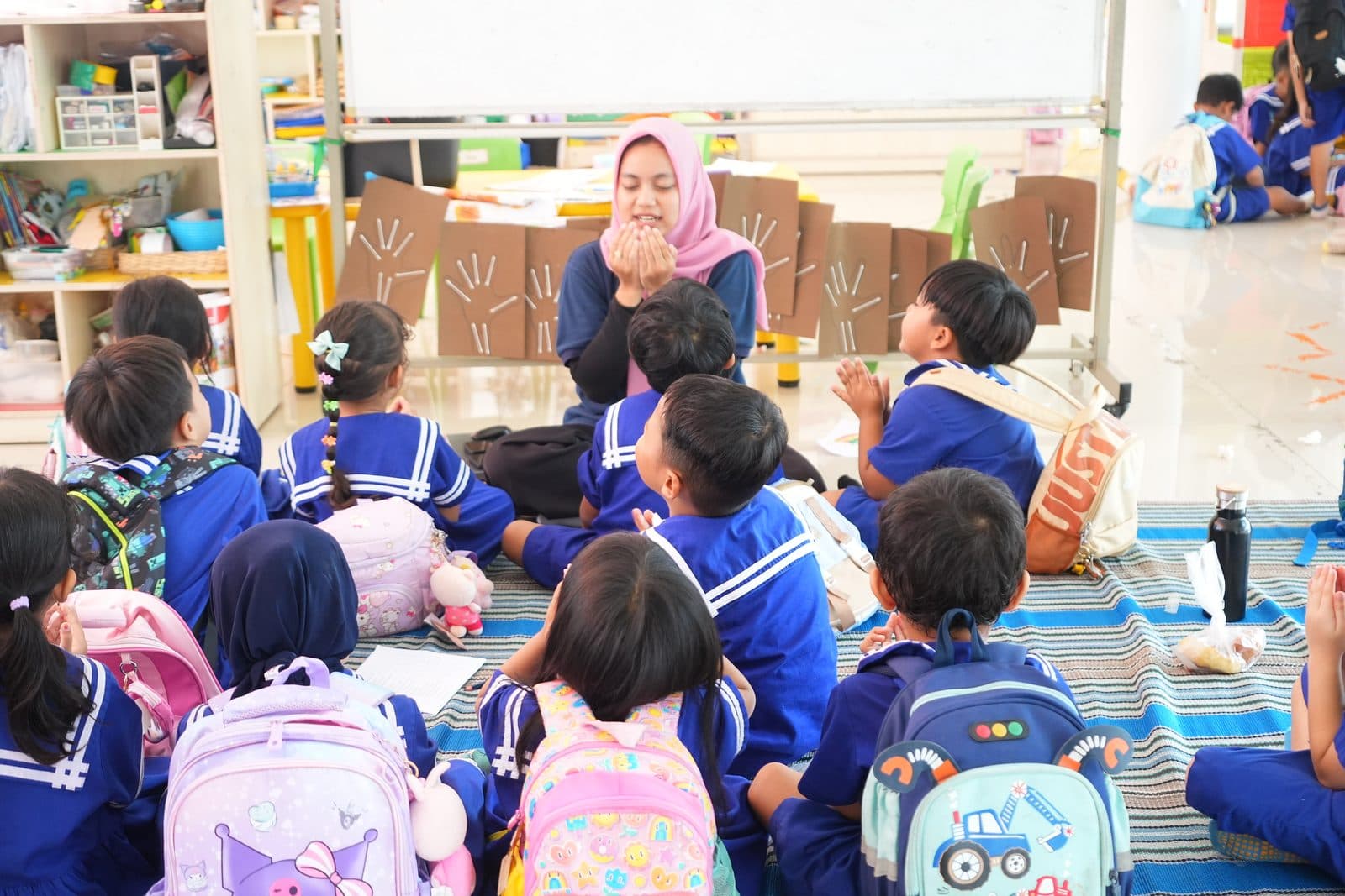 Children in blue uniforms sitting on a mat around their teacher